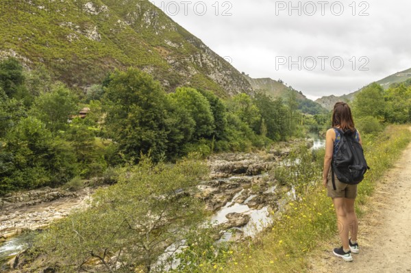 Female tourist with backpack walking along a path next to the sella river, contemplating the landscape in asturias, spain, during a cloudy summer day
