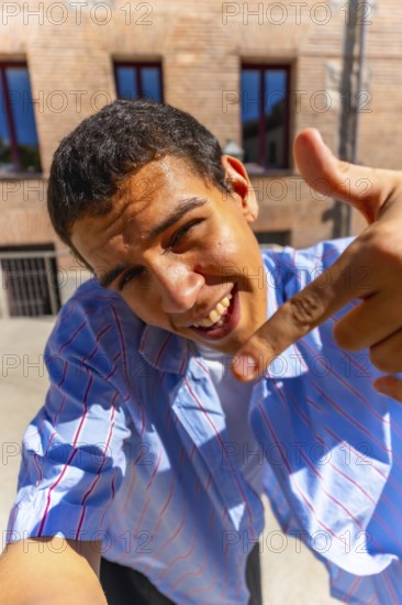 Young man smiling into camera and pointing down in a bright outdoor street selfie, casual striped blue shirt, confident upbeat vibe for vlogs, lifestyle and social media use