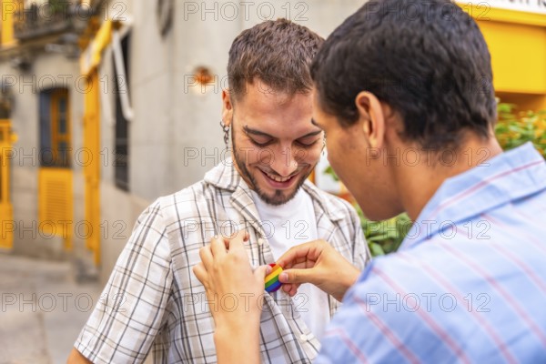 Young gay man gently attaching a rainbow pride flag pin to the plaid shirt of his smiling partner, celebrating love and lgbtq plus rights on a city street