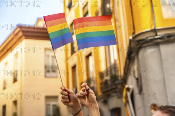 Hands are raising two small rainbow flags, symbolizing lgbtq plus pride, diversity, and equality, against an urban background during a daytime celebration or protest event