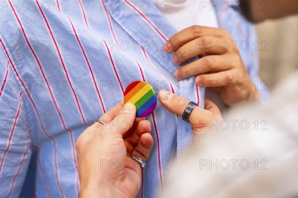 Young couples hands pinning a rainbow pride flag button to a striped shirt, close up symbolizing lgbtq plus support, love, equality, solidarity and human rights