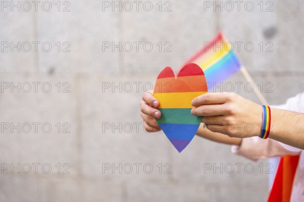 Person's hands holding a rainbow colored heart and a small pride flag, with a rainbow bracelet on the wrist, representing love, equality, and human rights