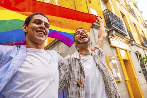 Happy young gay couple holding a rainbow flag and cheering loudly during a pride parade, expressing happiness and freedom against a vibrant city building