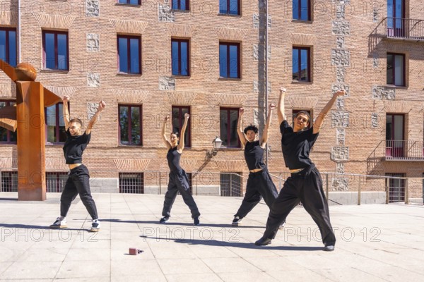 Four young male and female dancers performing a synchronized urban choreography with arms raised, expressing passion and energy against a brick building backdrop on a sunny day