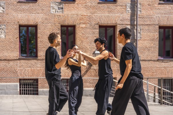 Group of diverse young performers wearing casual black clothing, happily dancing a contemporary routine together outdoors against a rustic brick building on a sunny day