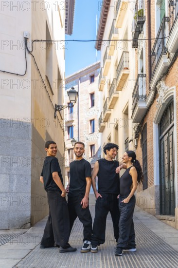 Group of diverse dancers wearing black casual clothing are posing and smiling while looking at the camera in a narrow city alley under a bright sky, symbolizing urban culture and friendship