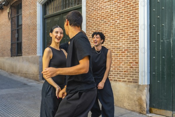 Young friends in black outfits laughing and dancing together on a city street by a brick wall, sharing joy, energy and playful urban togetherness in daylight