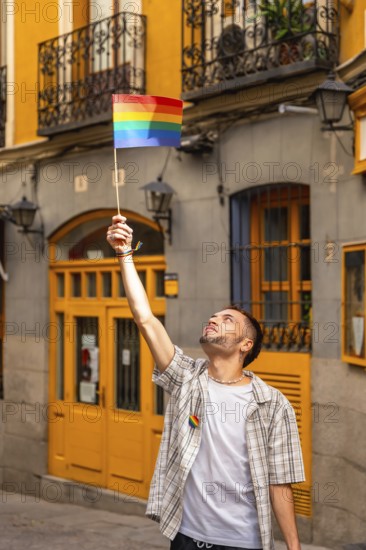 Young man standing in a city street, raising a small rainbow flag with arm outstretched, celebrating lgbtq plus pride, freedom, community and joyful support during daytime urban scene