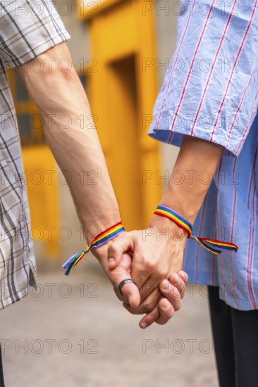 Male couple showing love and support for each other by holding hands with rainbow flag bracelets, symbolizing their lgbtq plus pride and relationship against a vibrant yellow background