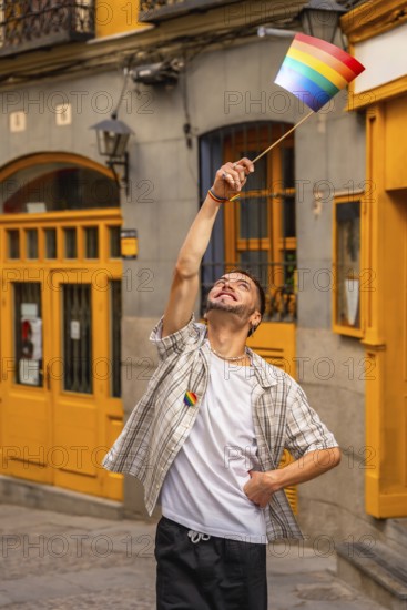 Young man standing on a street, smiling and raising a small rainbow flag, expressing happiness and support for the lgbtq community during a celebration event