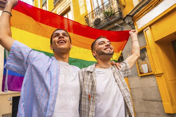 Happy young men joyfully embracing and holding a colorful rainbow flag together, looking up in a moment of celebration and pride on a vibrant city street