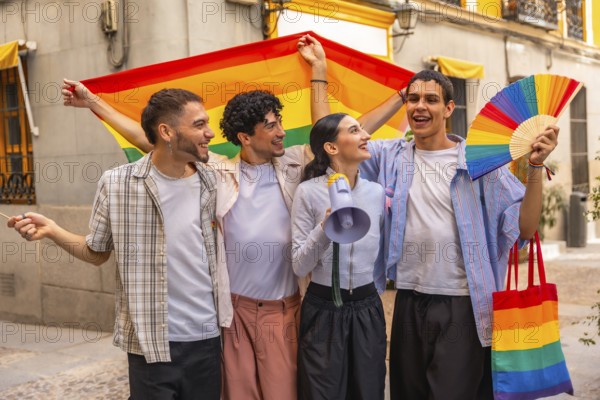 Diverse group of young adult friends smiling and holding a rainbow flag, fan, megaphone, and tote bag, celebrating pride and their community in an urban setting