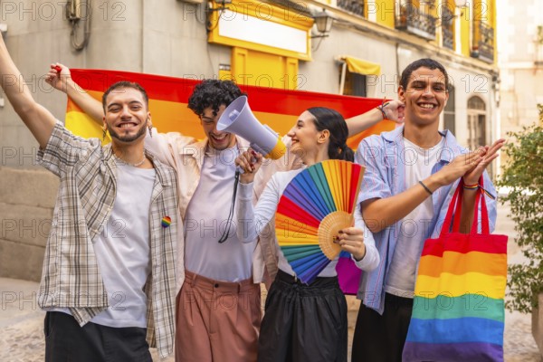 Diverse group of young friends standing together, holding a rainbow flag, fan, and megaphone, joyfully celebrating lgbt pride, diversity, and equality on a city street