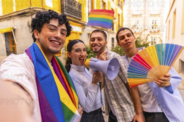 Group of diverse young people smiling and celebrating gay pride movement while holding a rainbow flag, a megaphone, and a colorful hand fan in a city street