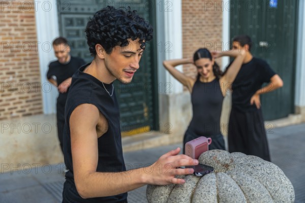 Young man playing music from a smartphone and portable speaker as urban dancers warm up on a city street, sharing rhythm, friendship and modern street culture