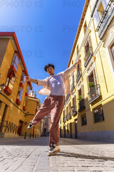 Young man actively posing and dancing on a cobblestone city street in historic european architecture under bright blue sunny skies, expressing joy and energy