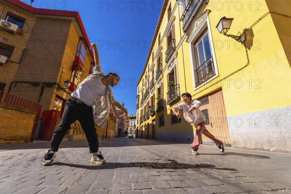Two young male dancers performing contemporary street dance move on a sunny cobblestone street, expressing dynamic motion and urban culture against yellow building facades