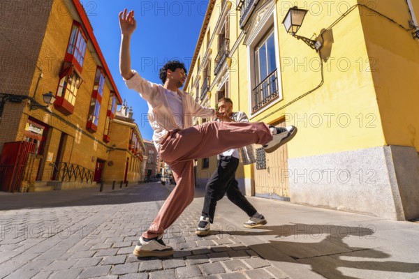 Two energetic young men are performing a dynamic dance routine on a cobblestone street, expressing happiness and freedom as bright sunlight illuminates the colorful urban architecture