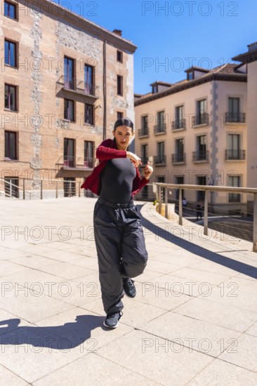 Young woman dancer performing dynamic contemporary routine on a sunny city street, showcasing urban fashion, confident expression and athletic movement against architecture