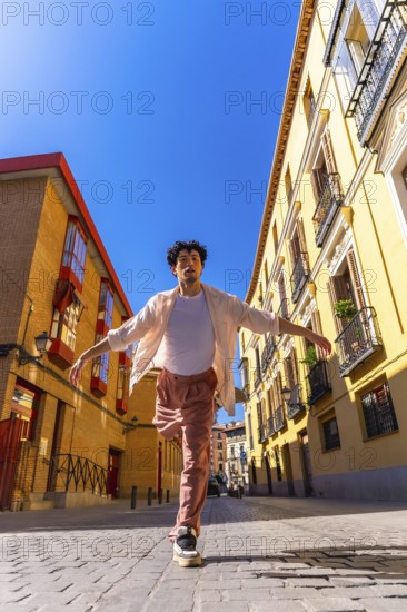 Young man actively dancing with outstretched arms on a sunny cobblestone street, expressing joy and freedom in a lively city environment with traditional buildings