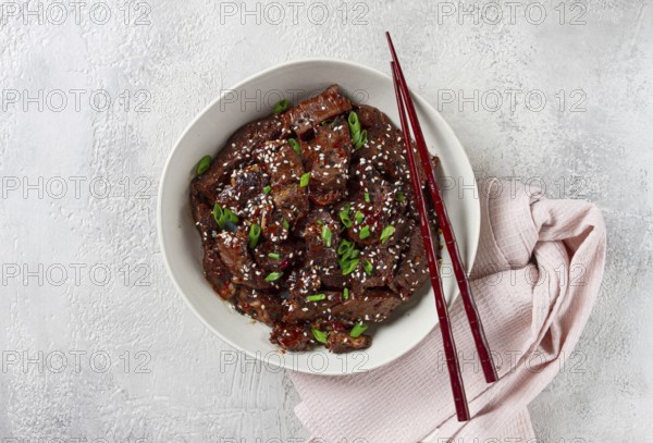 Crispy beef, with green onions and sesame seeds, in a bowl with chopsticks, on a textured surface, on a light background, no people