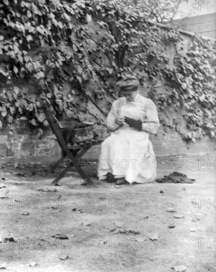Woman in domestic servant housemaid uniform sitting darning in garden setting, c 1900-1920