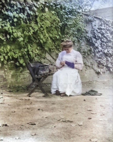 Woman in domestic servant housemaid uniform sitting darning in garden setting, c 1900-1920 colourised image