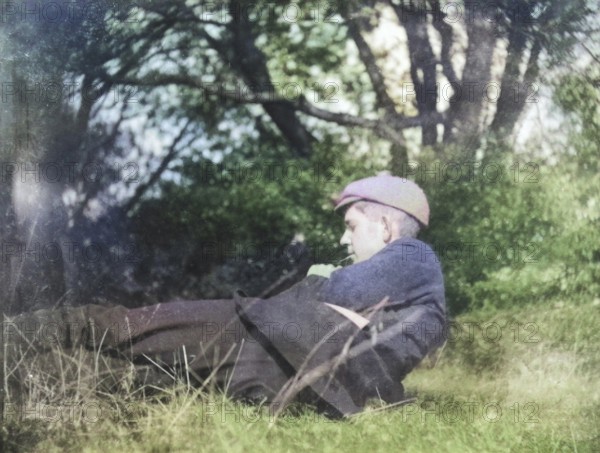 Colourised image of pensive young man lying in countryside field with trees c 1910-1920
