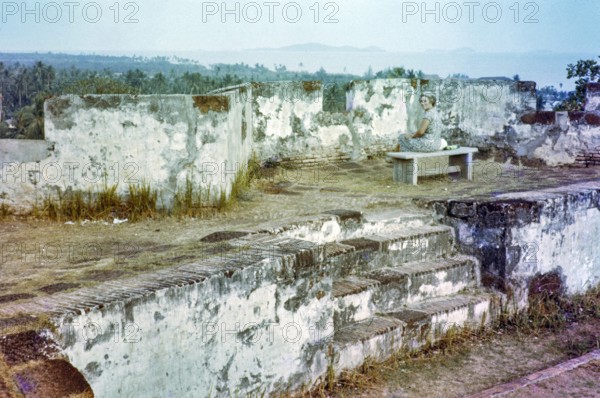 British expatriate tourist woman sitting on bench by walls ruin thought to be Malacca or Melaka, Malaya, Malaysia, south east Asia c 1964