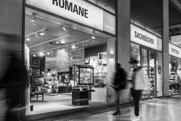 Black and white photograph, Dussmann GmbH, bookshop, urban scene in the evening with reflections in the window pane and a view of the displays and shelves, Berlin, Germany
