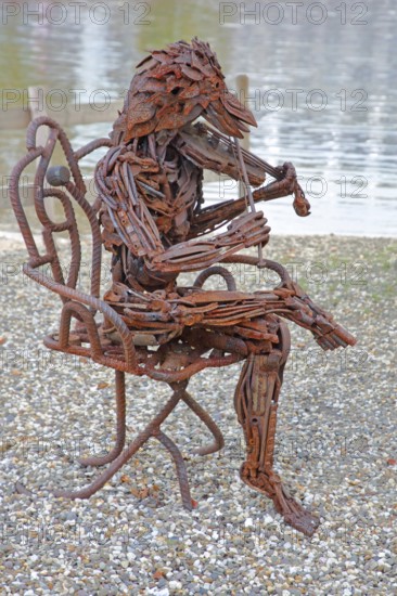 Sculpture The Concert by Günther Fahrner, rusty metal sculpture, sheet metal, modern art, bizarre whimsical musician sitting in a chair, violin player, bird figure, hybrid creature, playing music, pond, beach, shore, Bastinsweiher, Rhineland, Stolberg, North Rhine-Westphalia, Germany
