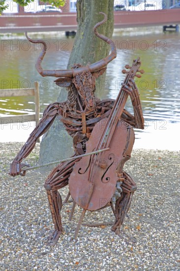Sculpture The Concert by Günther Fahrner, rusty metal sculpture, sheet metal, modern art, bizarre whimsical musician sitting in a chair, cello player, hybrid creature, playing music, pond, beach, shore, bull figure, Bastinsweiher, Rhineland, Stolberg, North Rhine-Westphalia, Germany