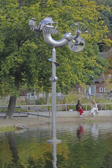 Sculpture Birdman by Albert Sous and two seated woman in conversation, people, pond, modern art, abstract art, stainless steel, metal sculpture, bizarre bizarre bird figure, mythical creature, hybrid creature, wings, flying, wheel, free-standing, Bastinsweiher, Rhineland, Stolberg, North Rhine-Westphalia, Germany