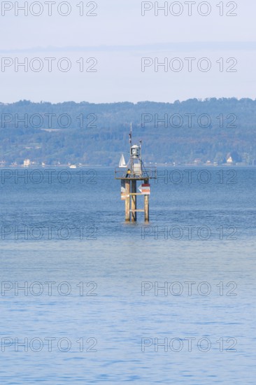 A lighthouse in blue water with a sailing ship, green hills and clear sky in the background, Constance, Lake Constance, Germany
