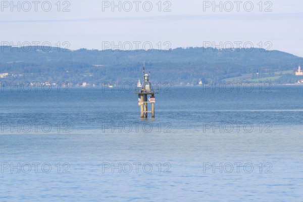 A tower stands in the calm waters of a large lake with green hills and a clear sky in the background, Constance, Lake Constance, Germany
