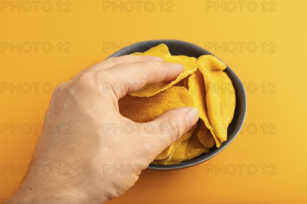 Dried Mango in blue ceramic bowl with hand on orange pastel paper background. Side view, close up. healthy food, minimalism