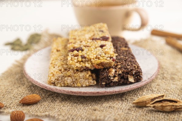 Granola bars with caramel, nuts, flakes in ceramic plate on white wooden background, beige linen napkin, cup of green tea. Side view, close up, selective focus