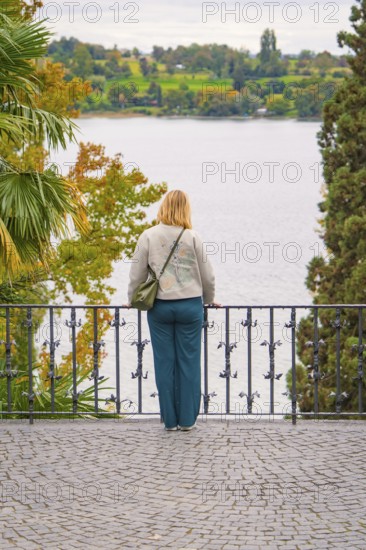 Person standing at the railing of a walkway and looking at a lake surrounded by trees in autumn, Mainau Island, Lake Constance, Germany