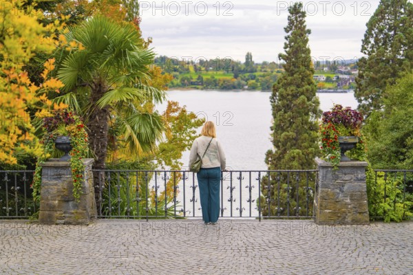 Person looking at a lake landscape with trees in warm autumn colours behind a railing, Mainau Island, Lake Constance, Germany