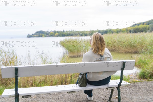 Woman on park bench looking at calm lake and autumn landscape, Mainau Island, Lake Constance, Germany