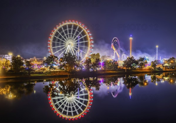 Folk festival in Stuttgart. Night shot with light effects. The 178th Cannstatter Volksfest on the Wasen is one of the most important traditional festivals in Germany. The Neckar River reflects the Ferris wheel and other attractions of the amusement park. Stuttgart, Baden-Württemberg, Germany