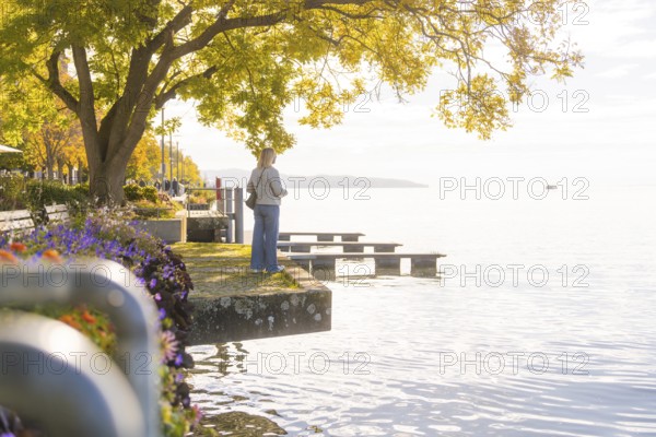 A woman stands on the lakeshore, surrounded by trees and flowers, while the scenery is bathed in warm sunlight, Überlingen, Lake Constance, Germany