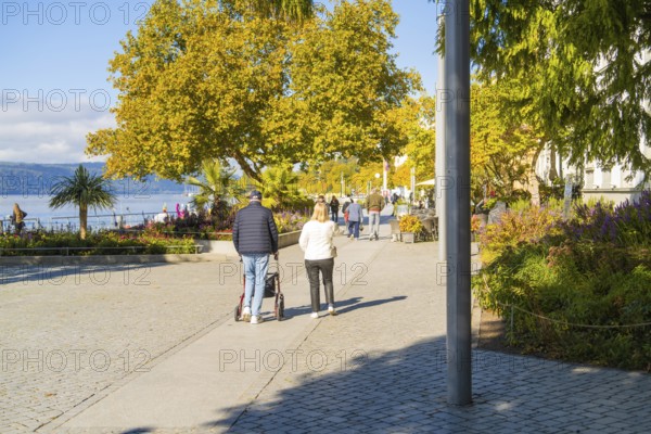 People walking on a sunny promenade along autumnal trees by the water, Überlingen, Lake Constance, Germany