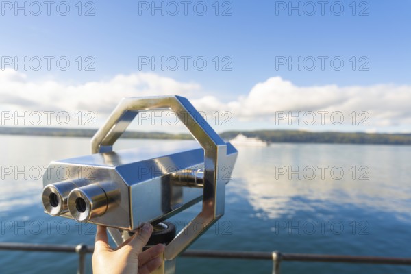 Hand holding binoculars on the lakeshore under a clear sky with clouds, Überlingen, Lake Constance, Germany