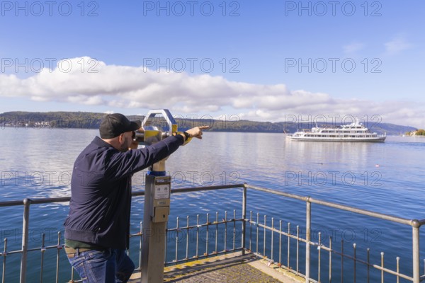 Man pointing with binoculars at a ship on a calm lake under a clear sky, Überlingen, Lake Constance, Germany