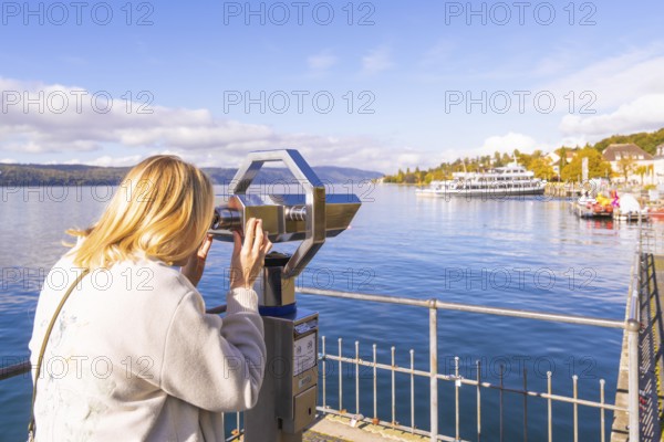 A woman looks through a telescope at a lake with ships and a clear autumn sky, Überlingen, Lake Constance, Germany