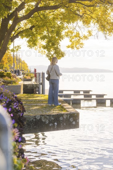 A woman stands on the shore of a lake in a relaxed atmosphere, surrounded by trees and flowers in the warm light, Überlingen, Lake Constance, Germany