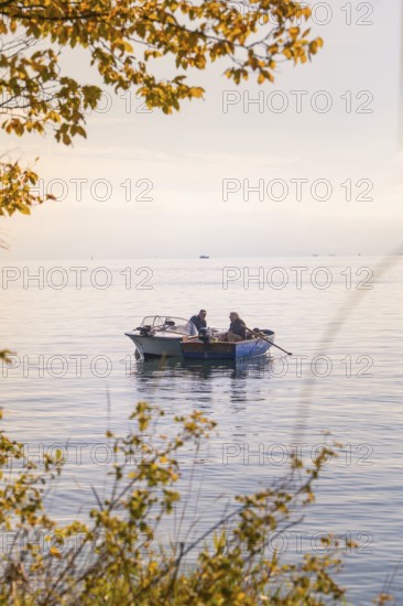 Two people fishing on a small boat in a calm lake surrounded by autumn leaves, Überlingen, Lake Constance, Germany