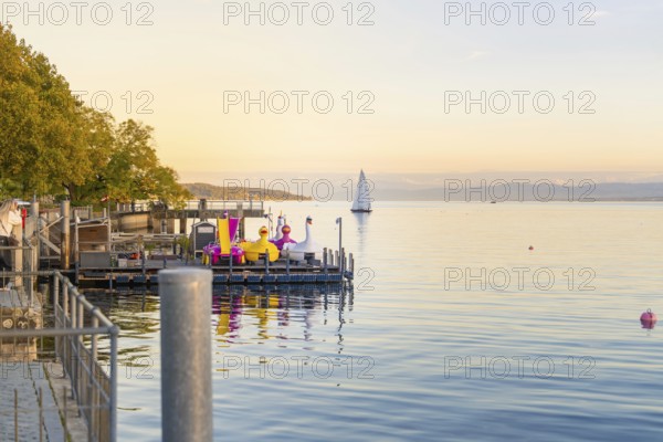 A river with colourful figures and a sailing boat on the calm lake at sunrise, Überlingen, Lake Constance, Germany