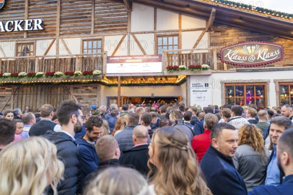 A large crowd waits outside in front of a festively decorated wooden house, Cannstatter Wasen fair, Stuttgart, Germany